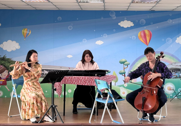 三位音樂家在舞臺上演奏。左側女性拉小提琴，中央女性彈鋼琴，右側男性演奏大提琴。背景為藍天白雲與熱氣球的卡通壁畫。舞臺布置溫暖童趣，呈現室內音樂會表演場景。