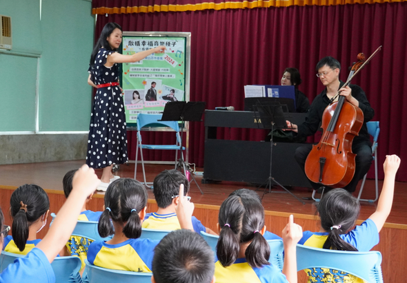 學校音樂教室內，一名女講師在舞台左側指著教育海報進行說明，舞台右側有鋼琴和大提琴演奏者。前景中坐著穿著黃藍色制服的小學生，舉手參與互動音樂教育活動。