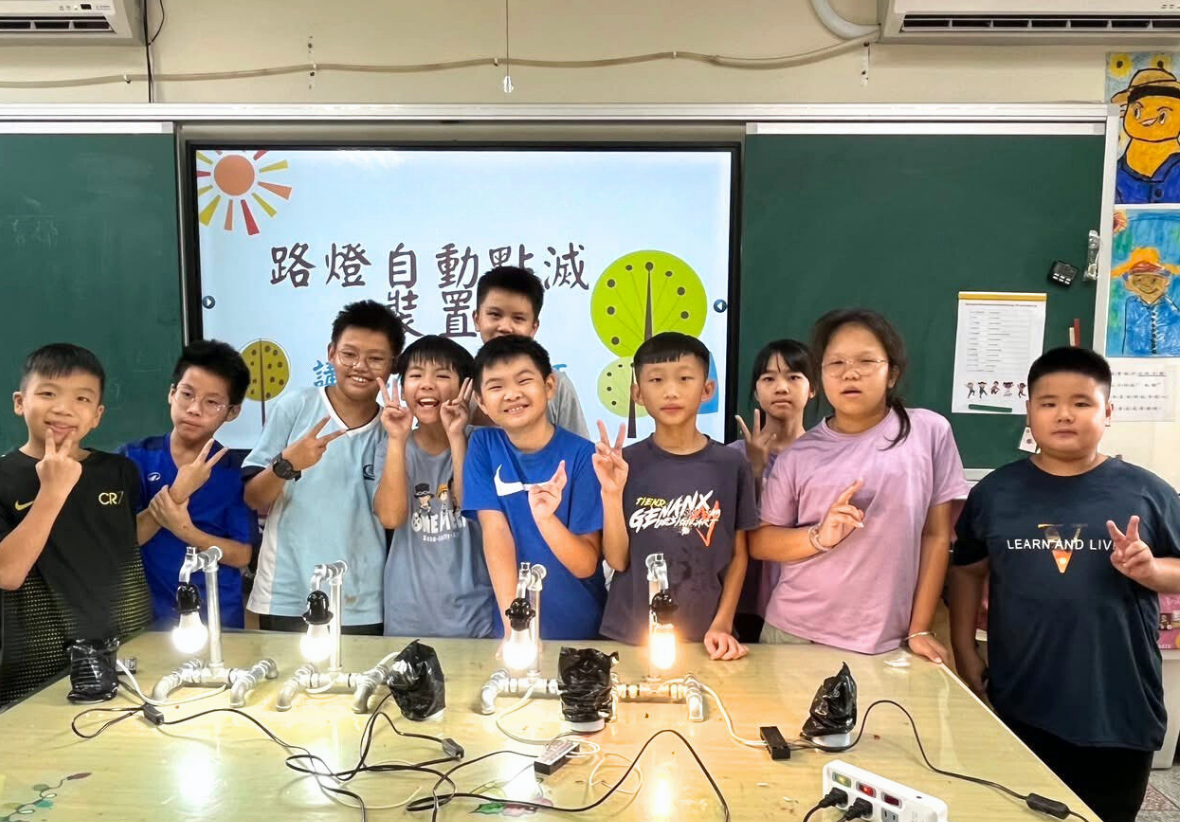 一群小學生在教室內進行科學實驗課程，桌上擺放多個點亮的燈泡裝置，展示路燈自動點滅的原理。學生們開心地擺出勝利手勢，身後投影幕顯示相關中文標題和綠色時鐘圖案。