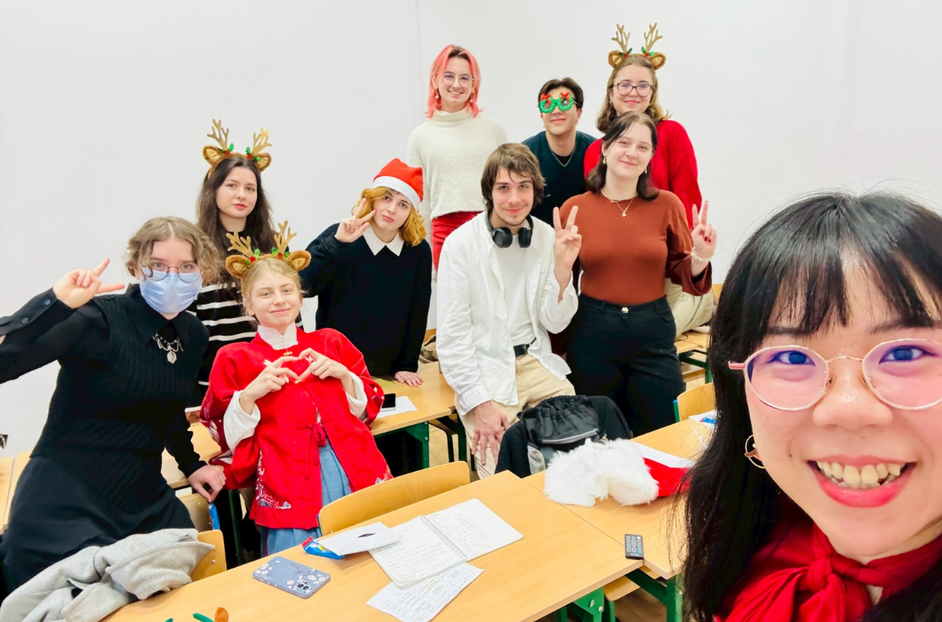 一群學生在教室裡進行聖誕節慶祝活動。他們戴著金色鹿角髮飾、聖誕帽和彩色眼鏡，擺出開心的姿勢。前方學生穿著紅色衣服，有人做愛心手勢，整個氛圍歡樂溫馨。