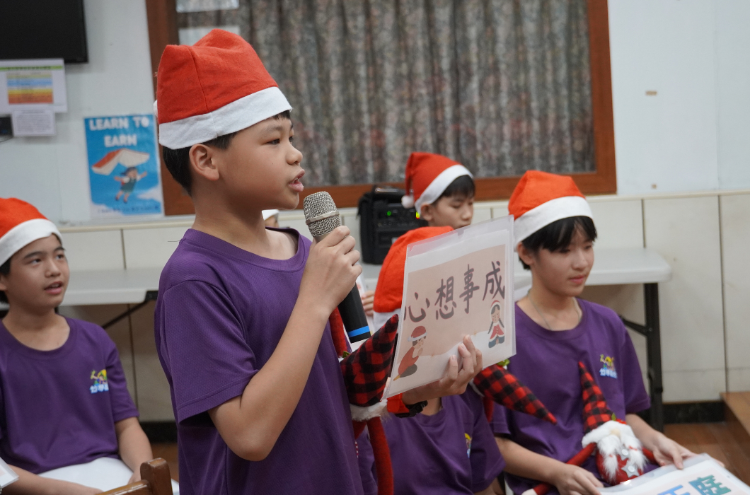小學生聖誕節活動現場，穿著紫色制服和聖誕帽的學童在教室內舉辦表演。前景中一名男童拿著麥克風發言，手持寫有中文字的卡片。其他學童穿著聖誕服裝坐在身旁，背景可見教室黑板和布置。