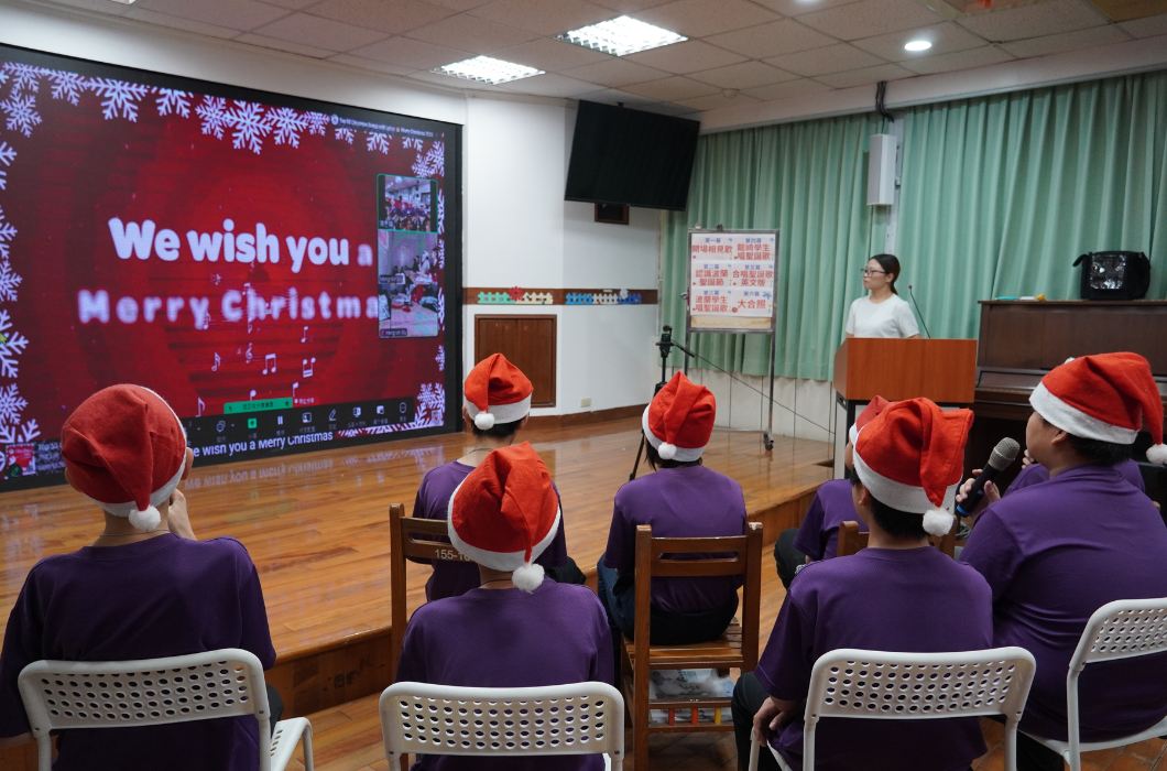 學校禮堂內，穿著紫色衣服、戴著聖誕帽的學生坐在椅子上參加聖誕慶祝活動。講台上一位老師主持活動，身後大螢幕顯示「We wish you a Merry Christmas」的聖誕祝福訊息。