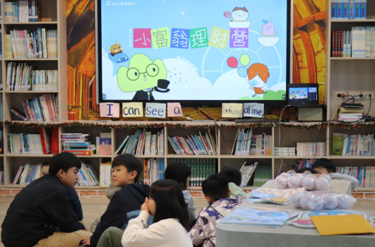 圖書館內，五位學生坐在地板上面向大螢幕觀看。螢幕播放著中文兒童教育內容，標題為「看看我們」，展示卡通人物和彩色圖案。背景為書架擺滿各類書籍。學生專注觀看教學節目。