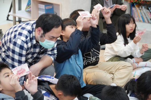 一位戴著口罩的男性教師在教室裡與多名學童互動，孩子們聚集在一起，有人舉起手中的卡片或教材。這是一場生動的教學活動，師生共同參與，充滿學習的氛圍。