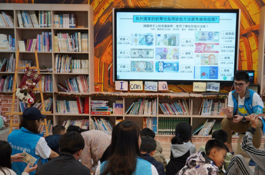 圖書館內一位講師向坐在地上的小學生們展示投影螢幕上關於鈔票防偽特徵的教育內容。螢幕顯示各種紙幣的安全防偽標記。背景是裝滿書籍的木製書架，學生們穿著藍色背心認真聆聽講解。