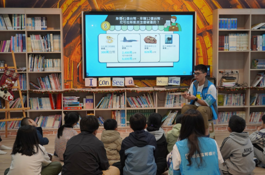 圖書館內，一位講師在大螢幕前向坐著的學生們進行教學演示。螢幕上顯示含有四個主題單元的課程內容。背景為書架，講師穿著藍色背心，學生們認真聽講。