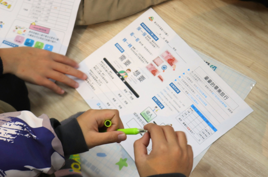 小孩在木質地板上用綠色螢光筆在學習教材上寫字，旁邊放著印有日文和圖表的練習紙，展示兒童進行教育活動的情景。