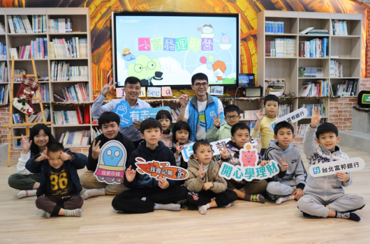 圖書館內，約十五名兒童坐在地上，兩位成人站在後方。背後螢幕展示彩色標題和卡通人物。兒童手持五彩繽紛的紙製標語牌，參加閱讀推廣活動。書架環繞四周。