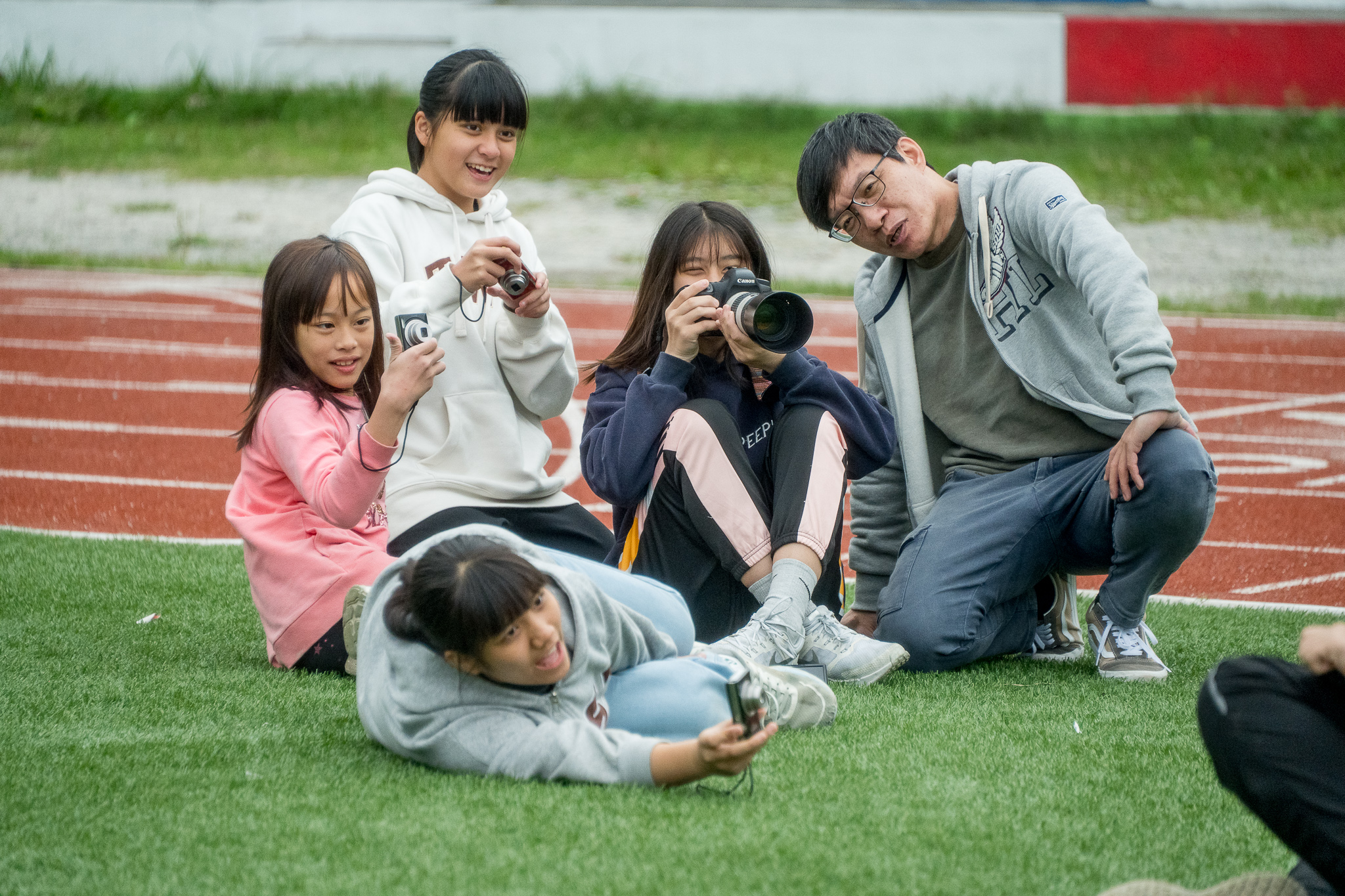 五位小學生在運動場上蹲坐成一圈，開心地舉起相機拍照。其中一人躺在草地上，其他人圍繞著她，有人舉著單眼相機、有人拿著小型數位相機，展現青春活力與友誼的溫馨場景。