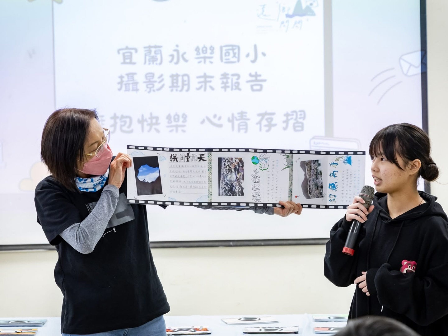 兩名學生在教室進行攝影期末報告發表。左側女學生展示電影膠捲形式的作品展示板，右側女學生拿著麥克風進行解說。身後投影幕顯示宜蘭水樂國小攝影期末報告相關文字。