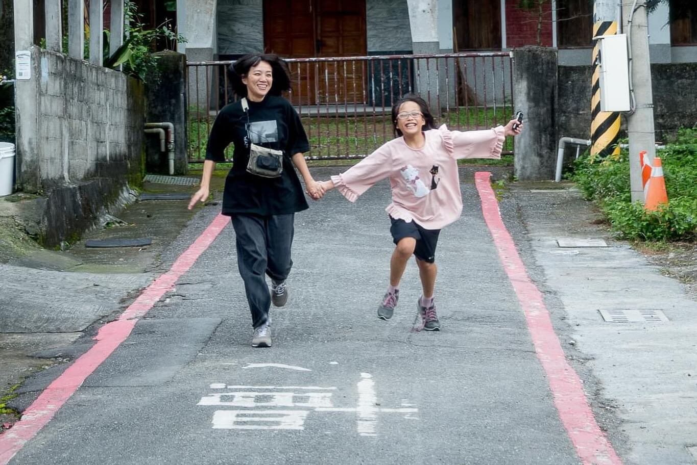 兩位女孩在街道上開心地奔跑，手牽著手，穿著運動服。街道兩側有粉紅色的跑道線標示，背景是混凝土建築和鐵製護欄。兩人都面帶笑容，展現青春活力。