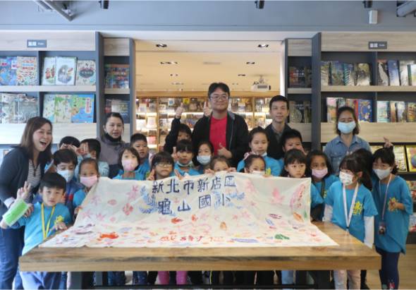 一群小學生與成人在圖書館內展示一幅彩繪布條，布條上寫有中文字。參與者們穿著藍色制服或衣著，大多戴著口罩，聚集在木製展示台前合影。背景為書架和展示櫃，場景呈現校園或社區藝術創作活動。