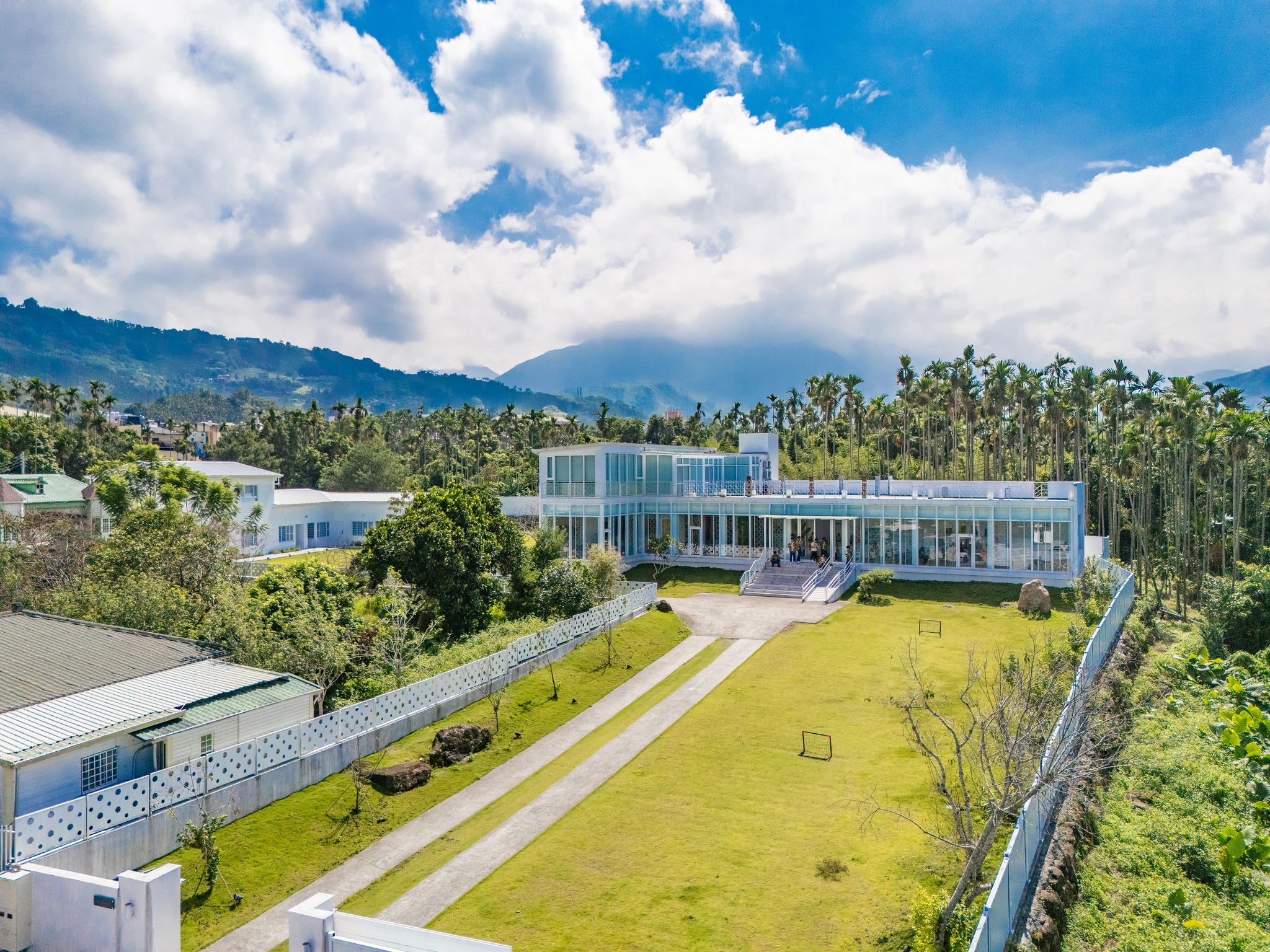 現代白色建築位於山谷綠地中，擁有大片玻璃窗和寬闊草坪。背景為棕櫚樹林與遠山，藍天白雲。建築周圍有白色圍籬和步行道，展現當代建築與自然景觀的融合。
