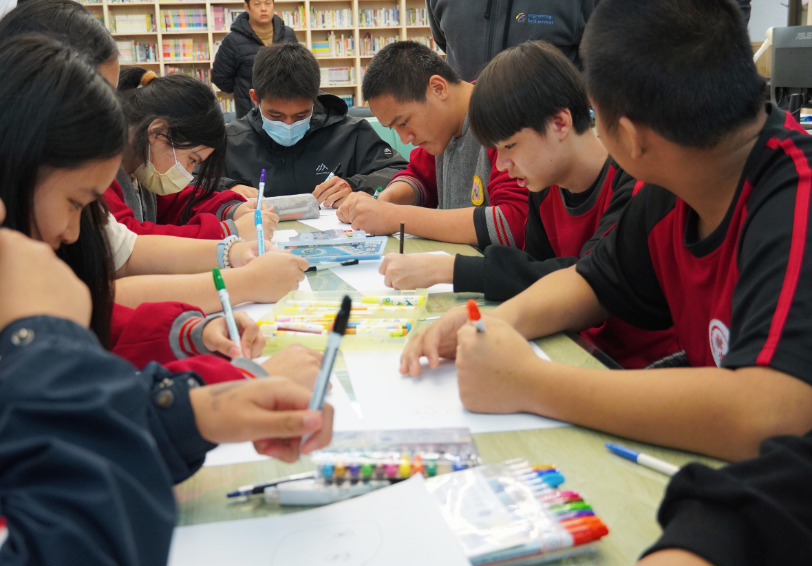 一群學生在圖書館內圍坐在桌邊進行美術創作活動。他們穿著紅黑色校服，拿著畫筆在紙張上繪畫。桌上擺放著彩色筆、顏料盤等美術用具。背景可見書架和指導老師，呈現熱烈的課堂學習氛圍。