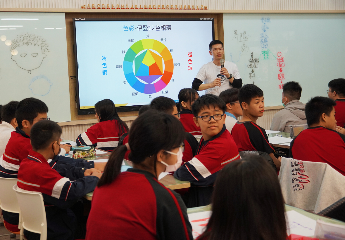 教室裡，一位男性教師站在觸控式電子白板前，手持麥克風向穿著紅色制服的學生講解色彩理論。白板上顯示「色彩-伊登12色相環」圖表，標示冷色調與暖色調。學生們坐在課桌前認真聽講。