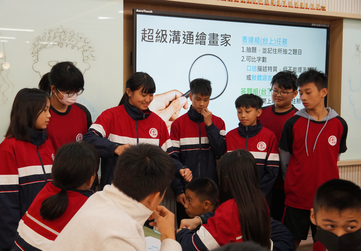 一群穿著紅藍色校服的學生圍成一圈，參與課堂活動。背後的觸控螢幕展示「超級溝通繪畫家」主題，包含抽題、可比字數等任務說明。學生們正專注地聆聽和參與討論，展現互動式學習的情景。