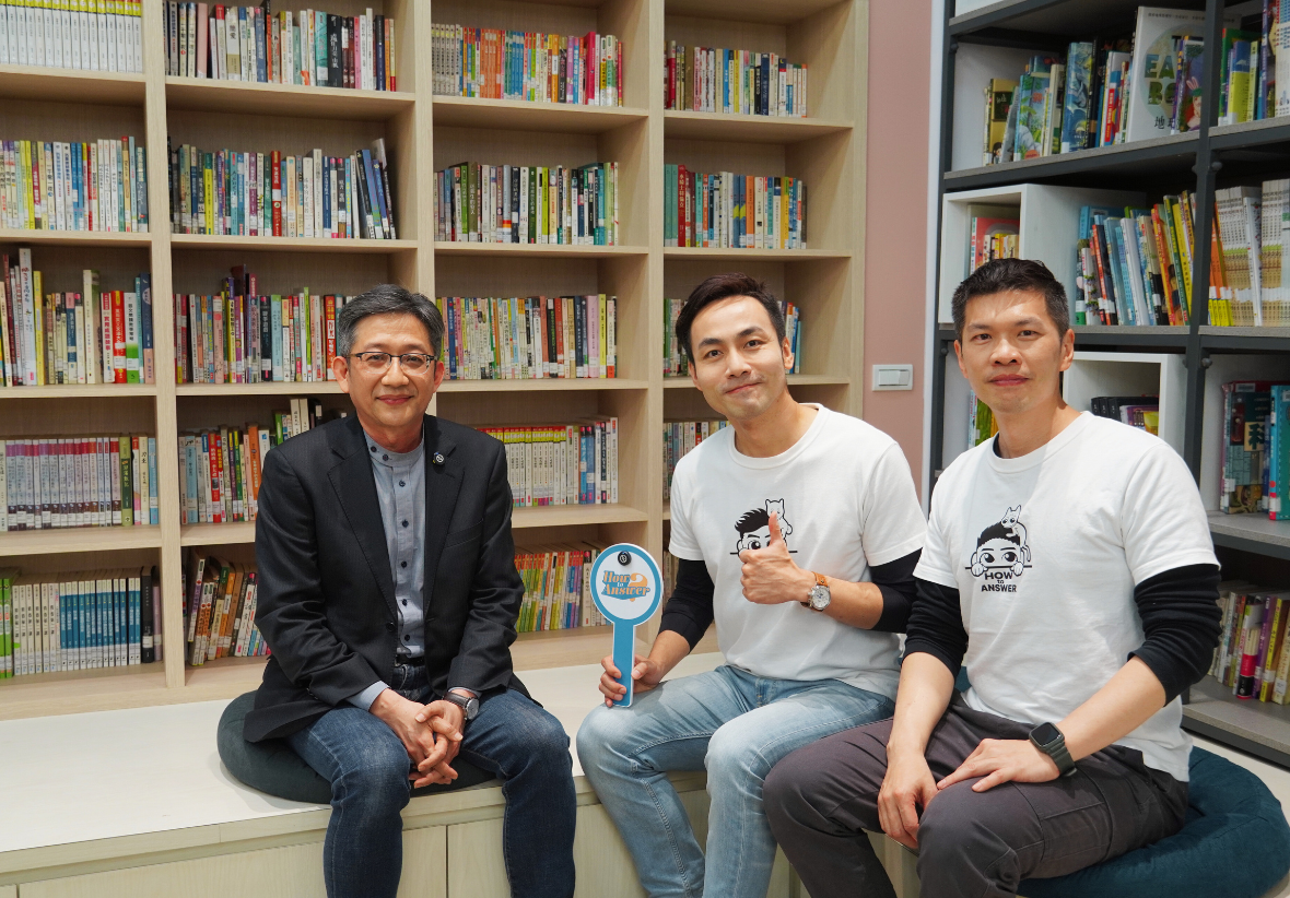 三位男性坐在圖書館書架前合照。左邊穿著黑色外套的男性，中間和右邊兩位穿著白色T恤。中間男性手持藍色圖書館標誌。身後是擺滿書籍的木製書架。