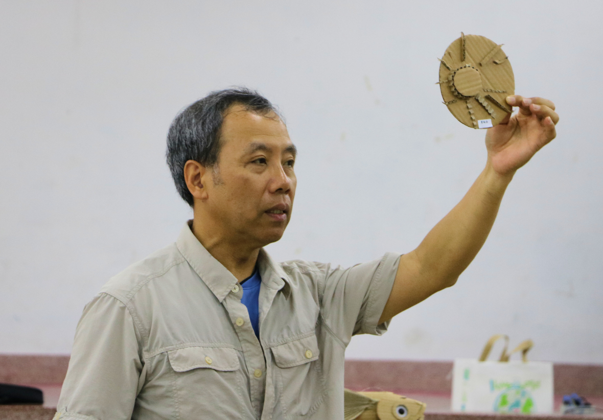 一位男性講者穿著米色工作服，舉起手中的竹編圓形工藝品，展示給觀眾。他在室內講堂進行教學或演示，身後可見傳統手工藝製品展示。