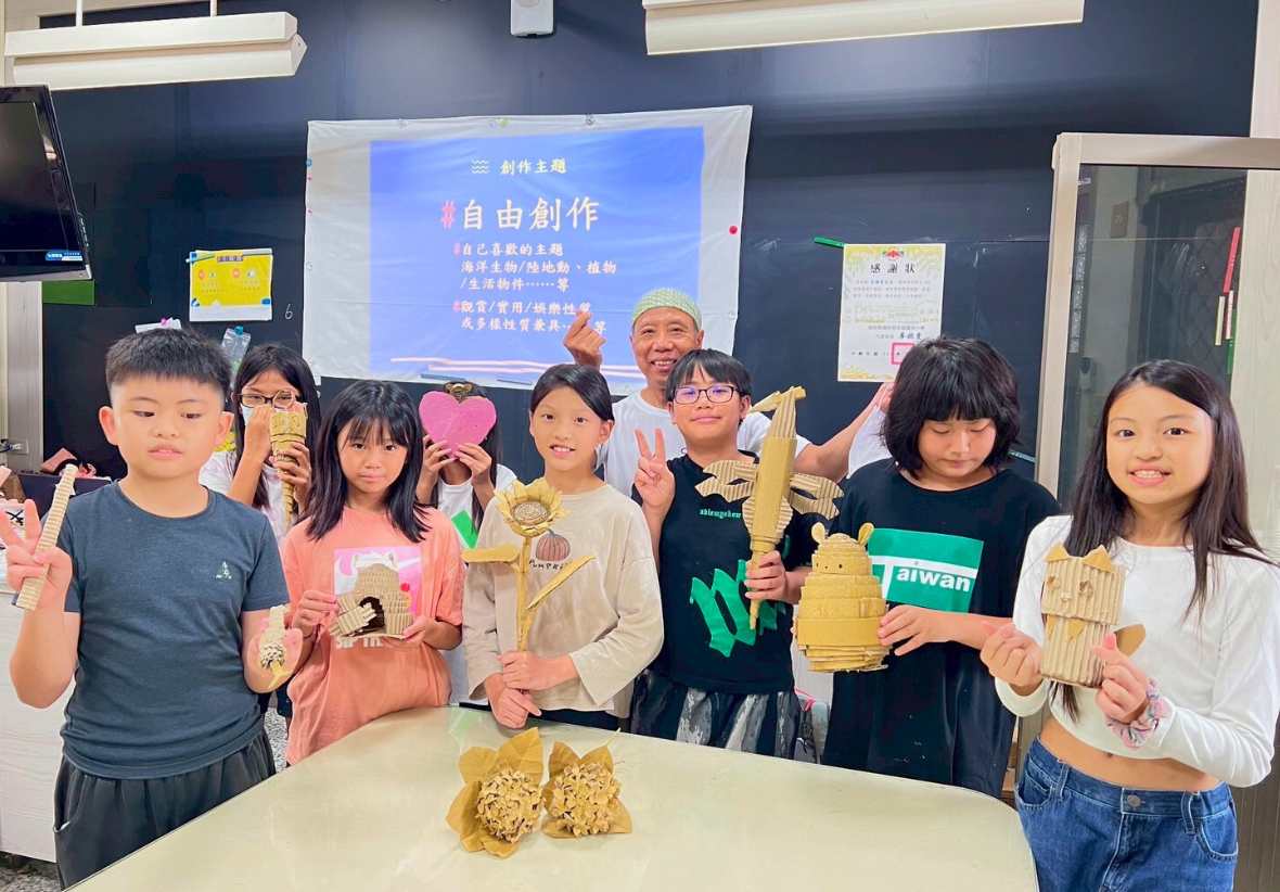 教室內，約十名學生展示自己用竹子創作的手工藝品，包括動物造型、建築模型等。背後投影幕顯示「自由創作」主題，講師站在中央。桌上放置完成的竹編作品。這是一場創意手工課程的成果展示。
