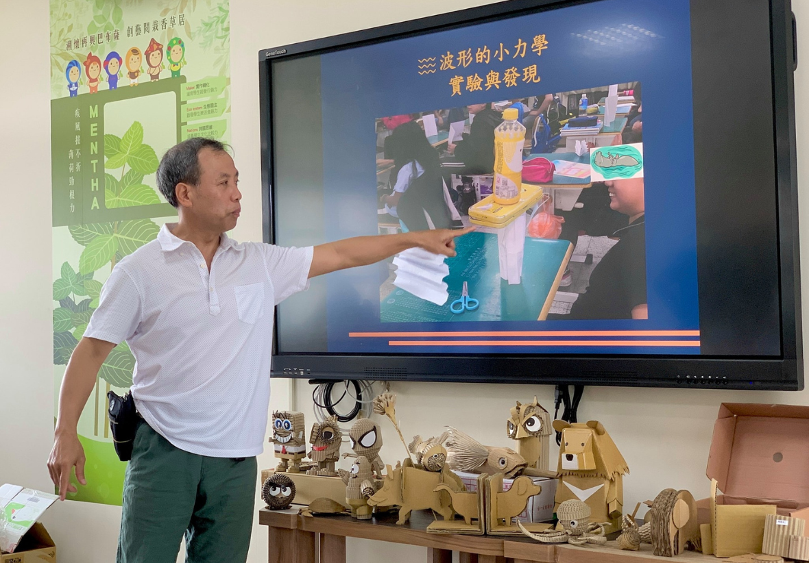 男性講師在教室內指著投影螢幕進行科學教學，螢幕顯示「波形的小力學 實驗與發現」主題，展示學生進行實驗的照片。講桌上擺放多個木製工藝品裝飾。左側牆上貼有綠色主題的教育海報。