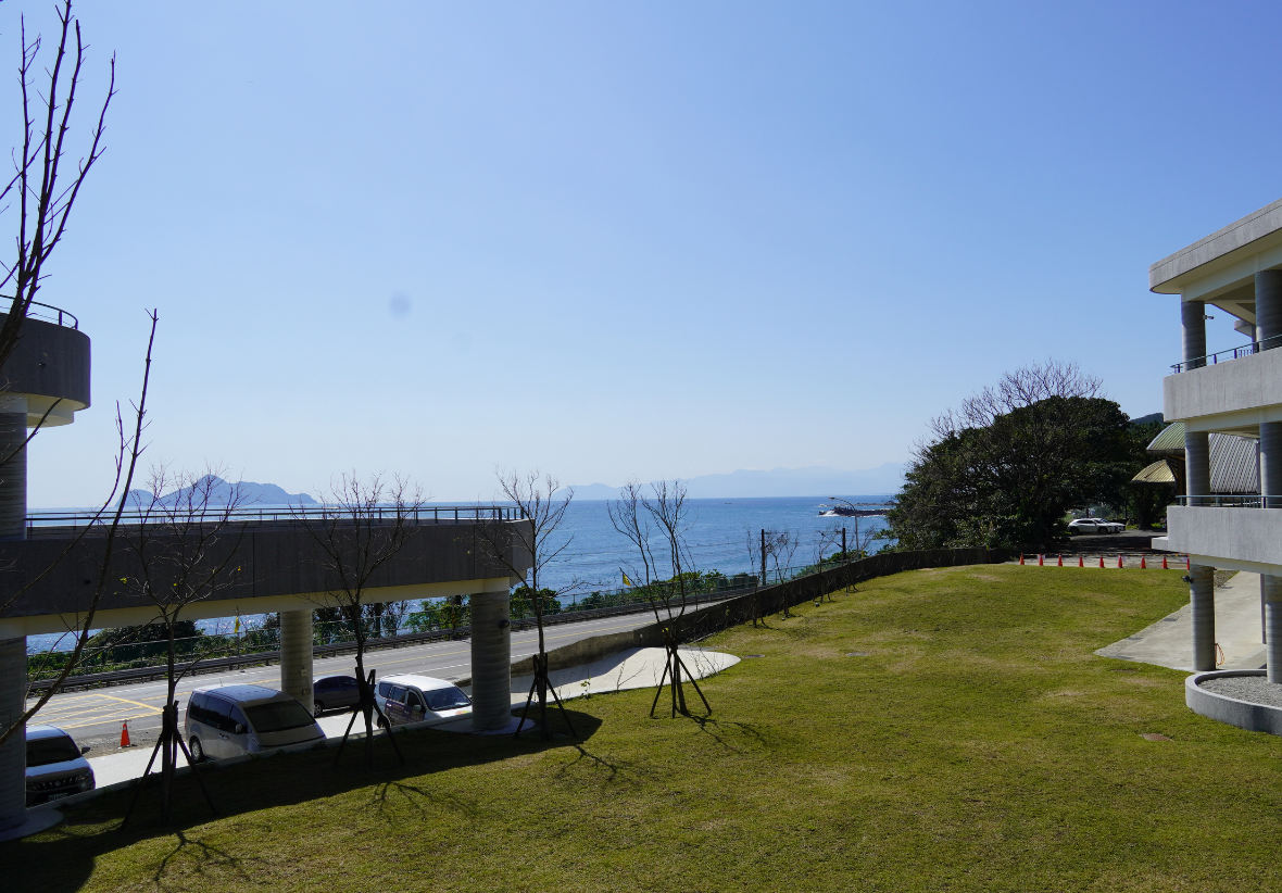 這張圖片呈現大里國小的校園戶外景致，包含圖片中央的草坪及停車車輛、右方的大里國小白色建築，以及遠方的大海及龜山島。