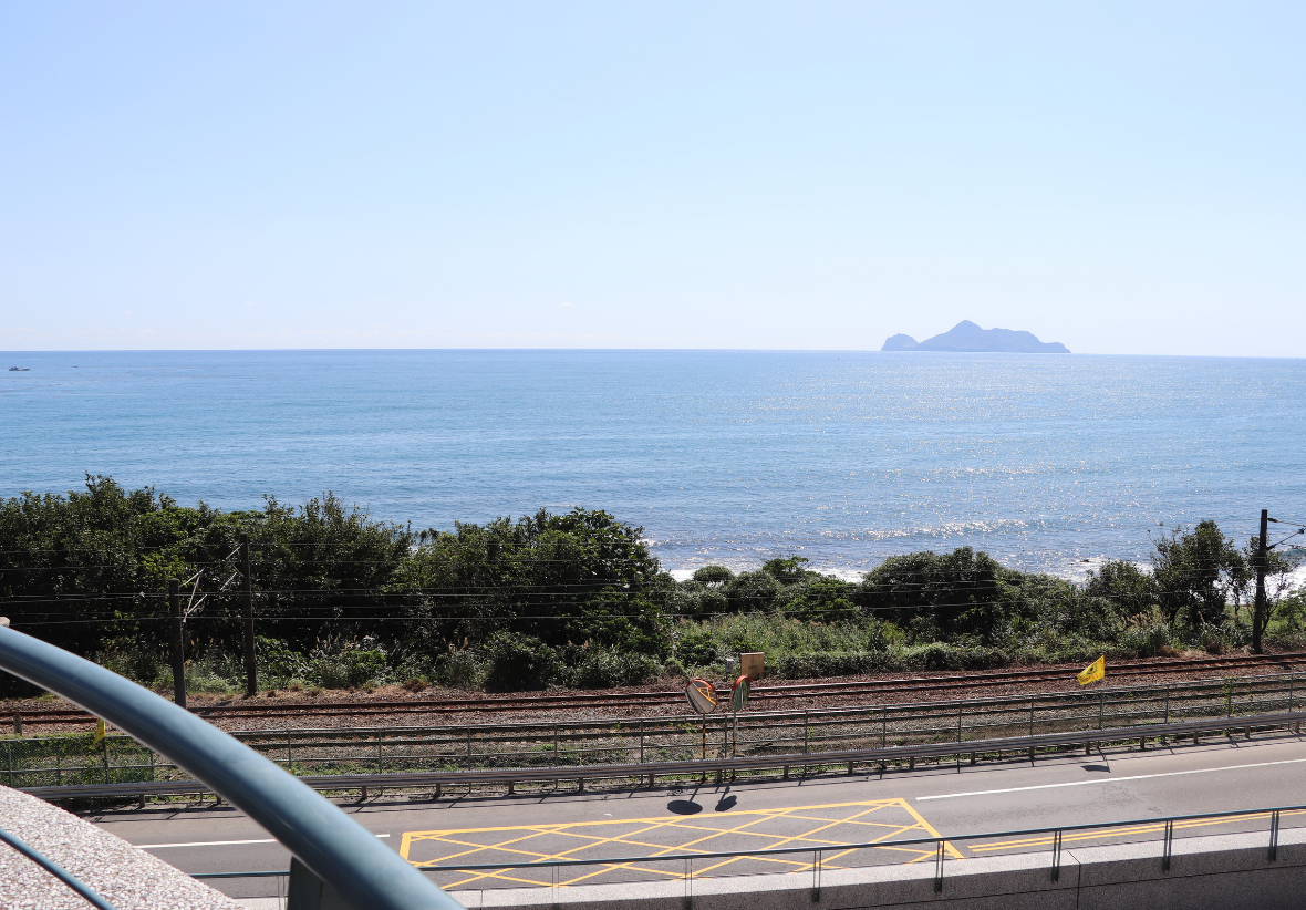 這是一張俯瞰海岸鐵道的照片。畫面由近到遠依序為馬路、鐵軌及綠色灌木叢。後方則是大海及龜山島 !