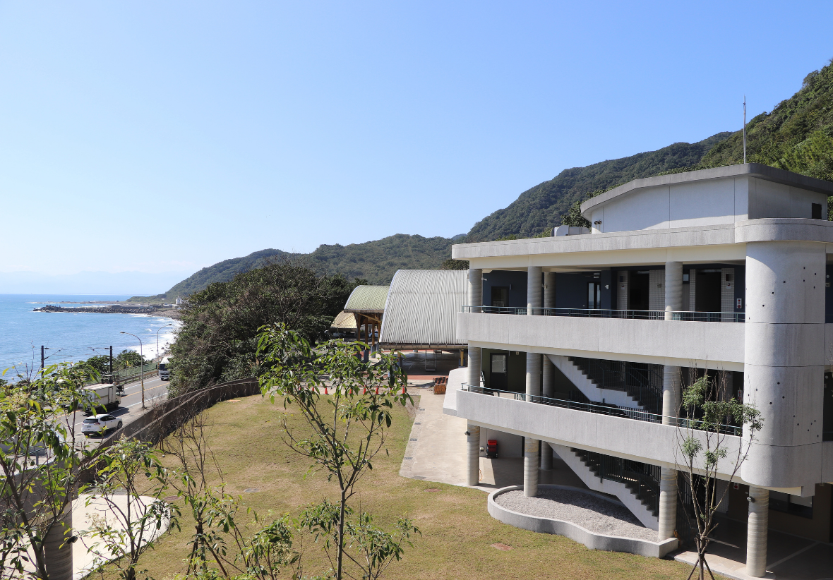 畫面主體是一棟三層樓高的白色校園建築，依山傍海，前方有綠色草坪與樹木，遠景則是連綿的山脈與海岸線 !