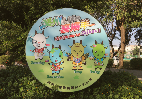 圖片為一個圓形的看板，標題為「溪州 Life 喜揚羊」。看板上有四隻造型各異的小羊吉祥物，分別代表學習、國際、創意與生態，底部寫著「我們都喜歡揚善助人的小羊」。