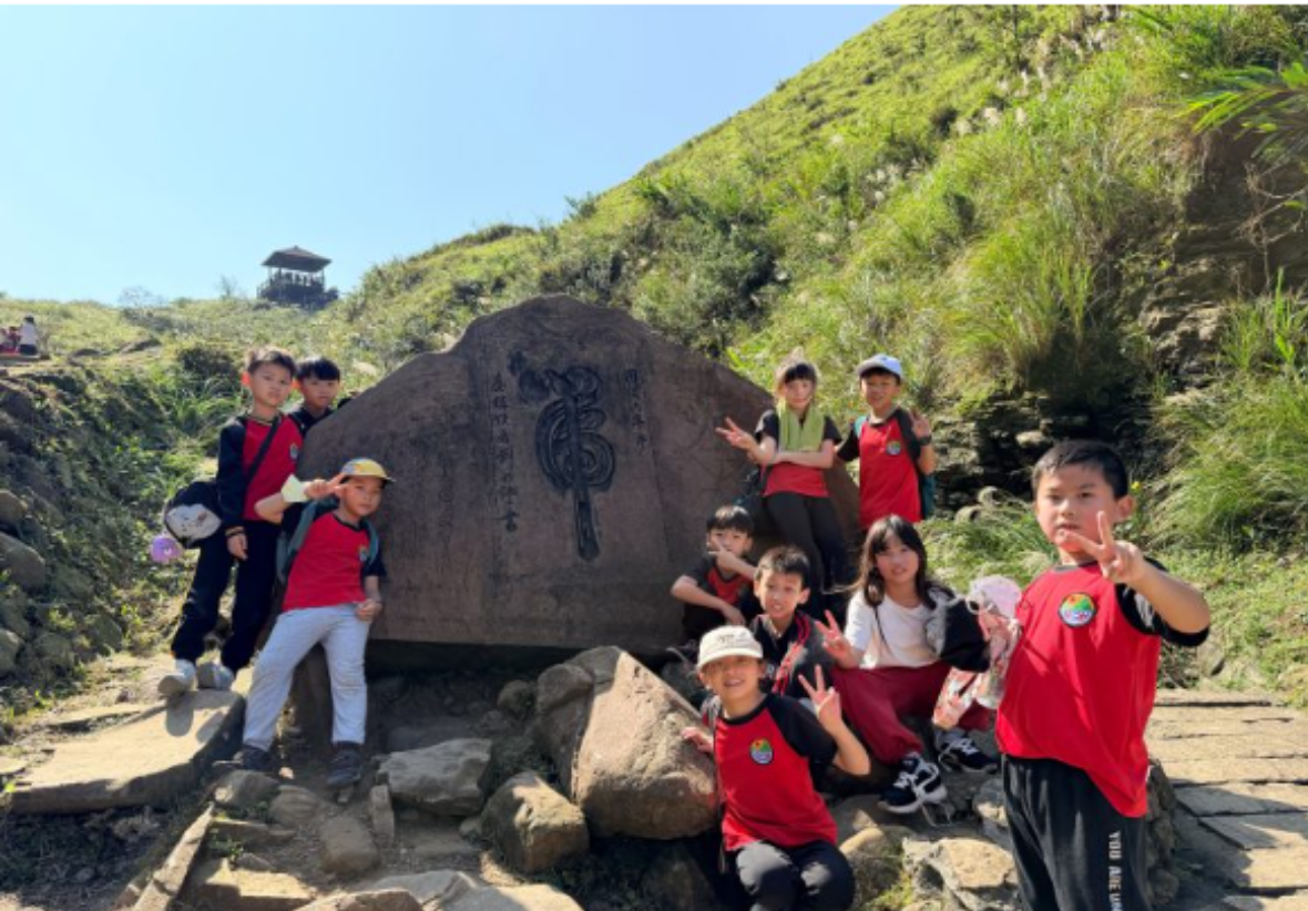 一群穿著紅色運動服的小學生在草嶺古道山徑步道上與石碑合照。背景為翠綠的山坡與藍天，展現戶外教學的歡樂氣氛。
