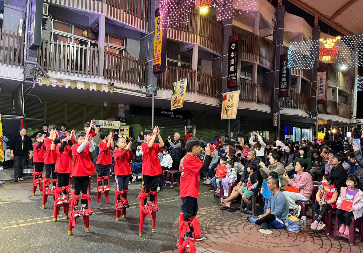 這是一場夜晚的戶外高蹺表演。一群穿著紅色運動服的孩子正在踩高蹺，吸引了大量民眾圍觀，展現了熱鬧的民俗技藝表演。