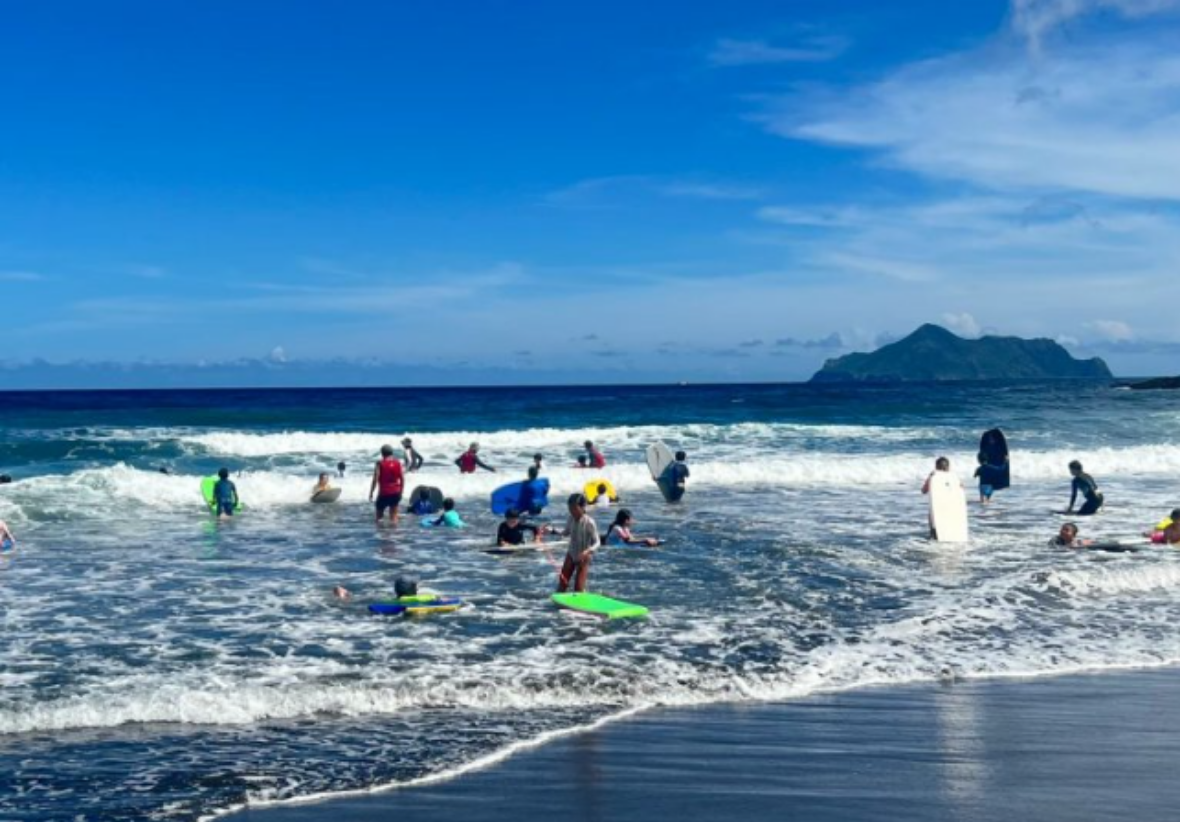 學生在湛藍的大海中體驗衝浪，海面上激起陣陣白浪，遠方可以看見清晰的龜山島輪廓。