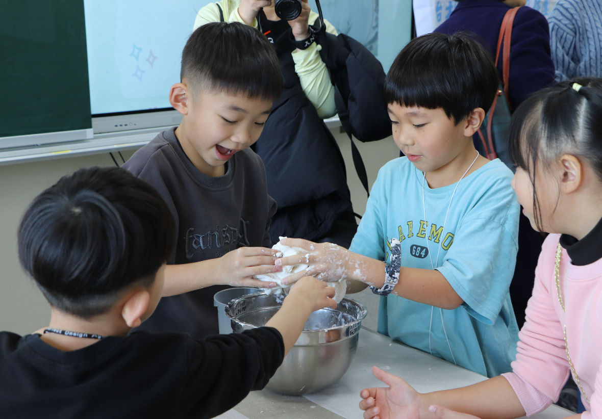 四位小學生圍在桌邊，正開心地一起揉捏麵團。桌上放著不鏽鋼盆，孩子們神情專注且愉快。