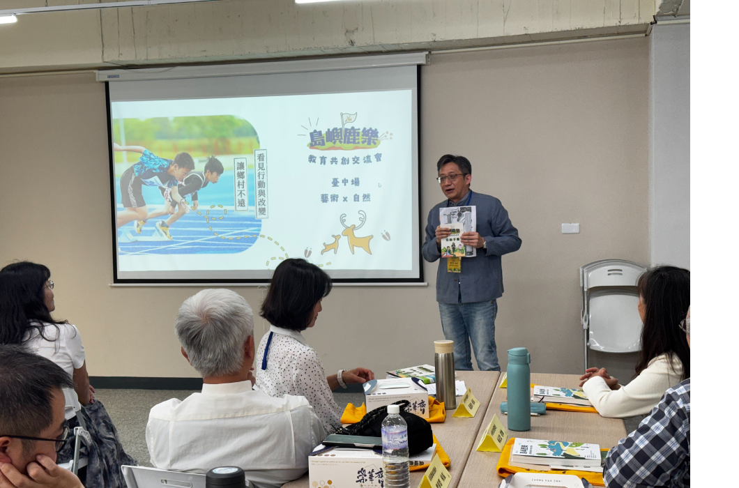 男性講者於室內分享教育經驗並展示書籍，後方投影「島嶼鹿樂 - 教育共創交流會_臺中場_藝術X自然」簡報畫面，參與者圍坐聆聽與討論。
