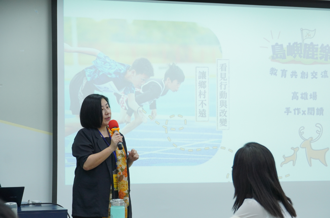 一位女性講師站在投影幕前，手持麥克風進行分享，螢幕顯示「島嶼鹿樂 - 教育共創交流會_高雄場_手作 X 閱讀」活動資訊。台下聽眾專注聆聽，呈現出教育交流座談的專業氛圍。