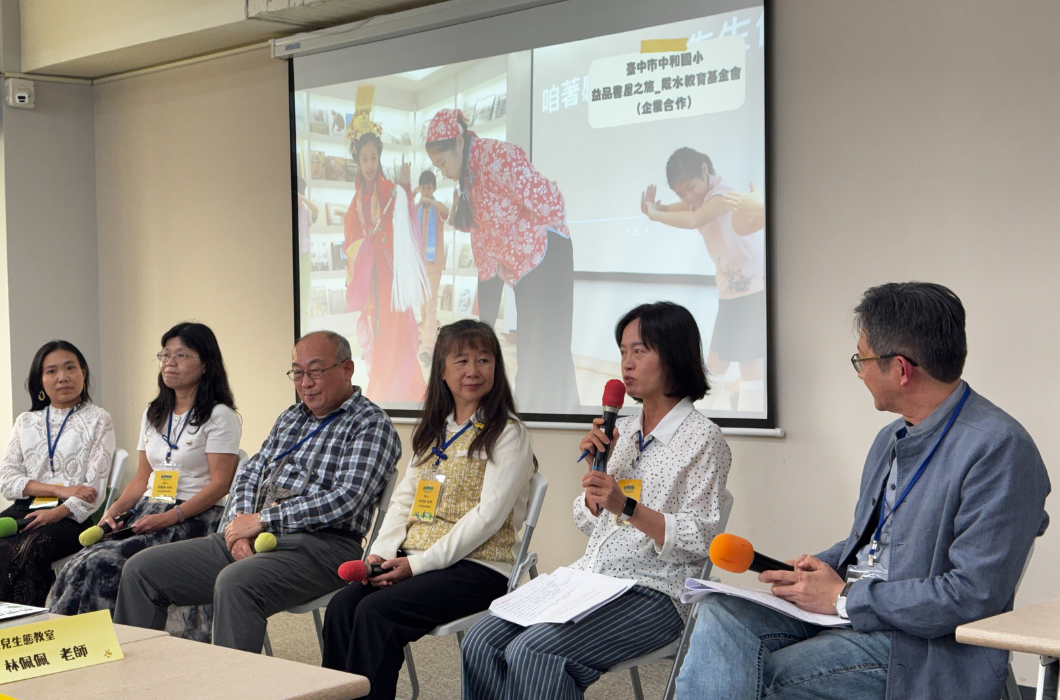 多位講者坐於教室前方進行座談交流，主持人手持麥克風發言，後方投影幕展示活動照片，現場氣氛專注熱絡。
