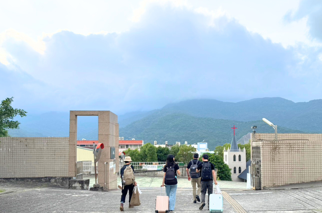 三位鹿樂平臺夥伴及一位計畫主持老師拖著行李走向校園出口，遠方可見山巒、雲層與教堂景色，畫面寧靜開闊。