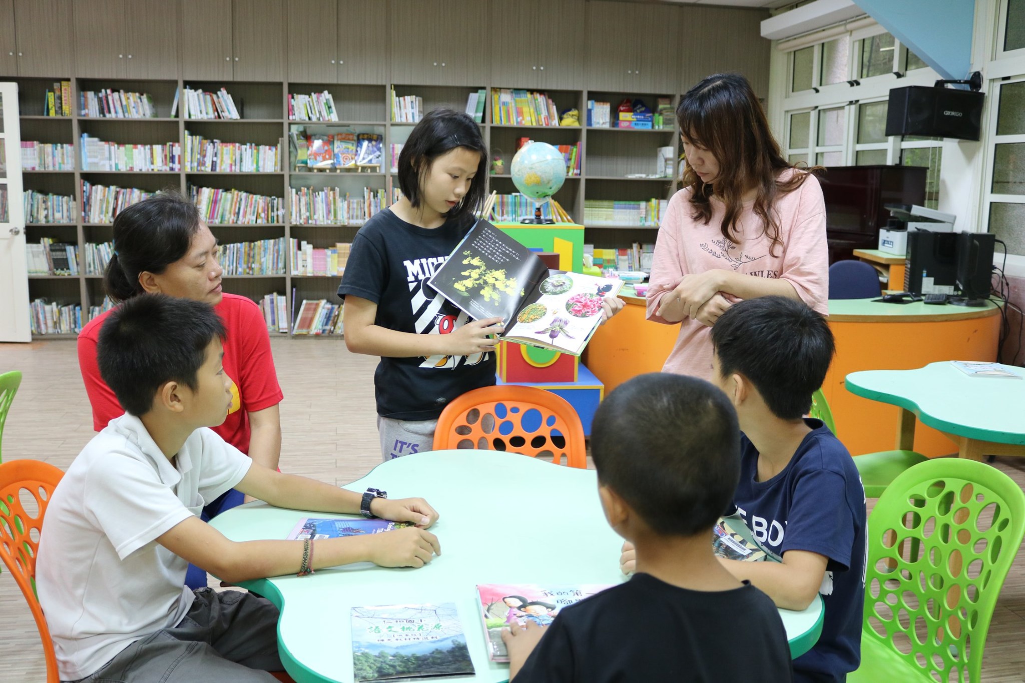 圖書館內的閱讀活動。一名女童站在淺綠色四葉草造型的桌子前，手裡拿著一本打開的繪本，正向坐在桌邊的其他學童們展示內容。旁邊有兩位大人陪伴，孩子們都專心地看著繪本。背景是排滿書籍的圖書館木製書架。