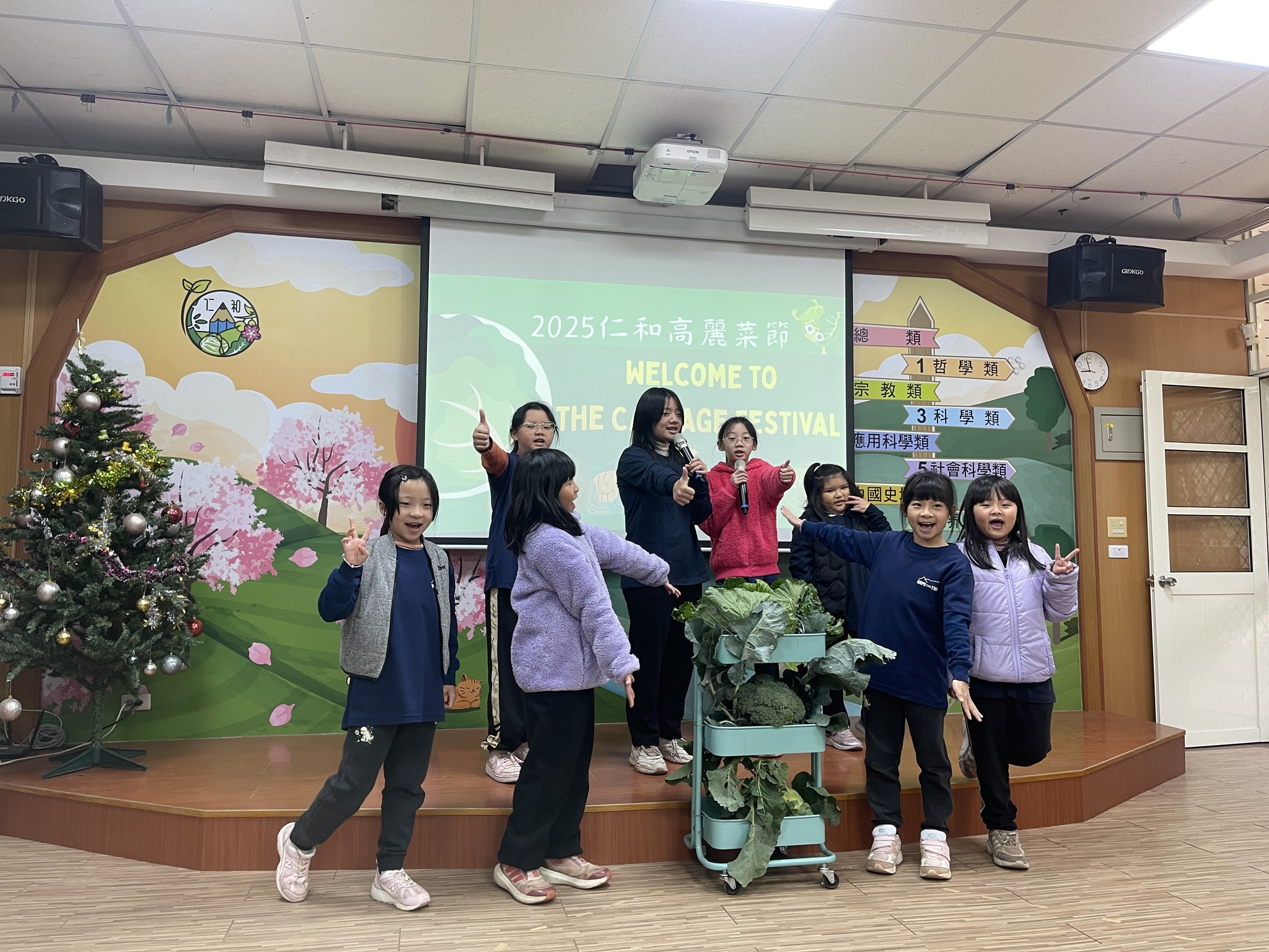 七名女童在室內舞台上充滿活力地表演。後方的投影布幕上顯示著「2025仁和高麗菜節 WELCOME TO THE CABBAGE FESTIVAL」。女孩們對著鏡頭開心微笑並擺出各種手勢，其中兩位手持麥克風。她們的身旁有一台裝滿巨大高麗菜的三層淺綠色推車。舞台左側擺放著一棵裝飾過的聖誕樹。