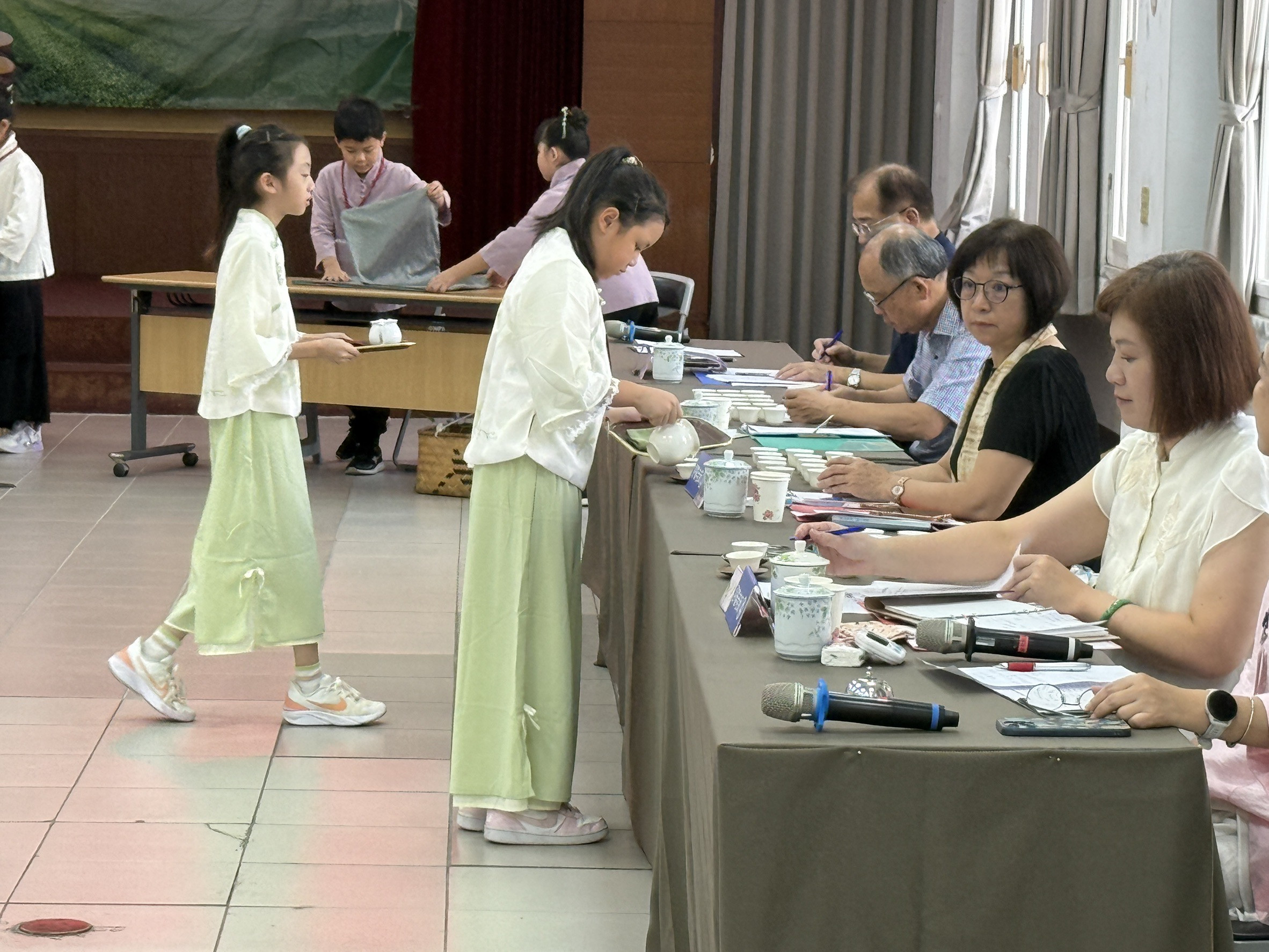 室內舉辦的茶道展演或檢定活動。兩位穿著淺綠色搭配白色傳統服飾的女童，正在長桌前進行泡茶。前方的女童正專注地拿著茶壺將茶水倒入茶杯中。長桌的另一側坐著一排大人（評審或老師），他們桌前擺放著許多茶具與麥克風，正一邊觀看學童的動作一邊在桌上的紙張做評分或紀錄。