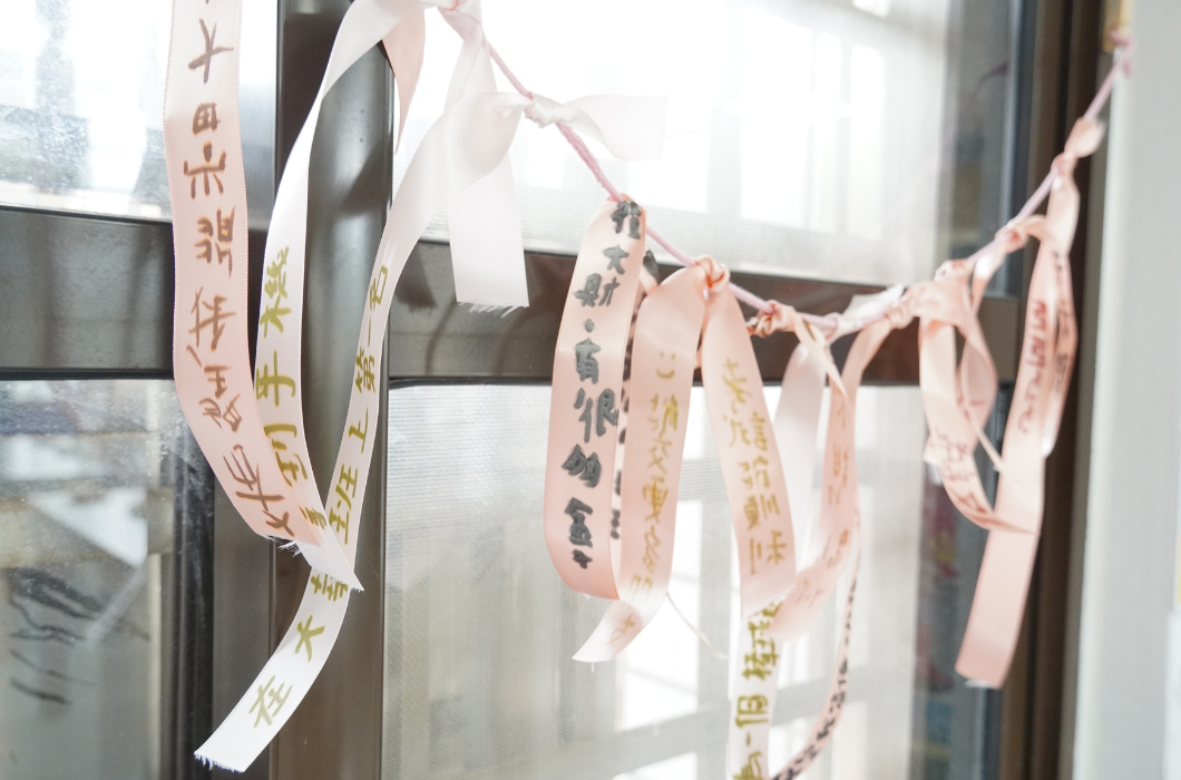 掛在窗戶邊上的粉紅色絲帶裝飾，絲帶上用手寫字體寫著「拿到手機」、「班上第一名」、「發大財，有很多錢」等各式願望。