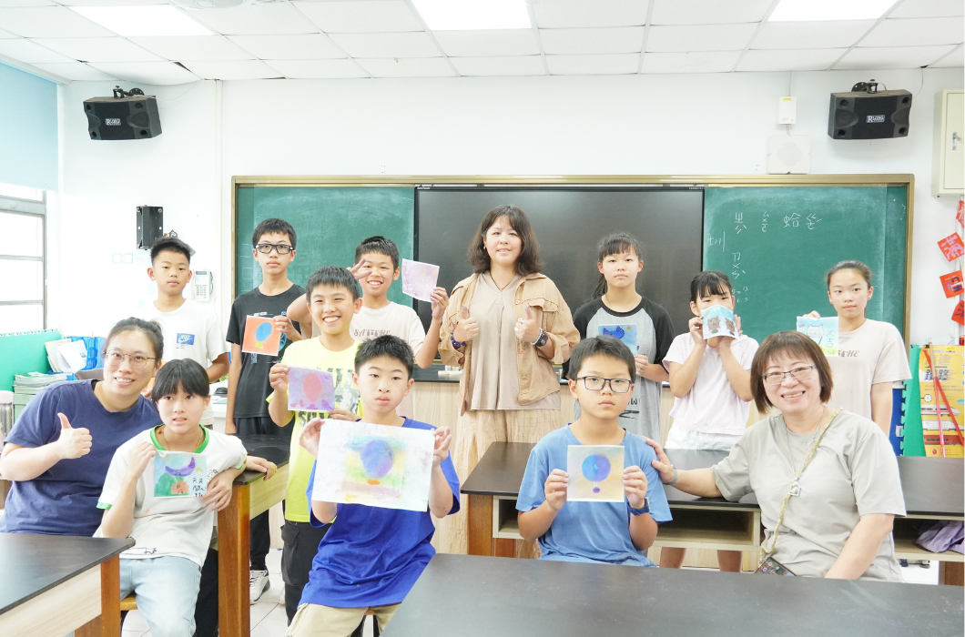 教室黑板前的大合照。多位學童高舉著自己的粉彩畫作，與三位女性指導老師及志工一起對著鏡頭開心微笑與比讚。