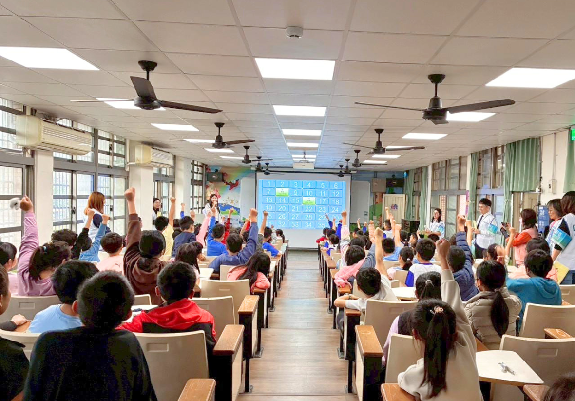 這是一張從階梯教室後方拍攝的大合照。教室內坐滿了國小學童，他們大多數都背對鏡頭，高舉雙手，積極參與台上的互動。投影螢幕上顯示著數字格子。幾位工作人員站在教室前方和兩側，微笑地看著學生，現場氣氛熱烈。
