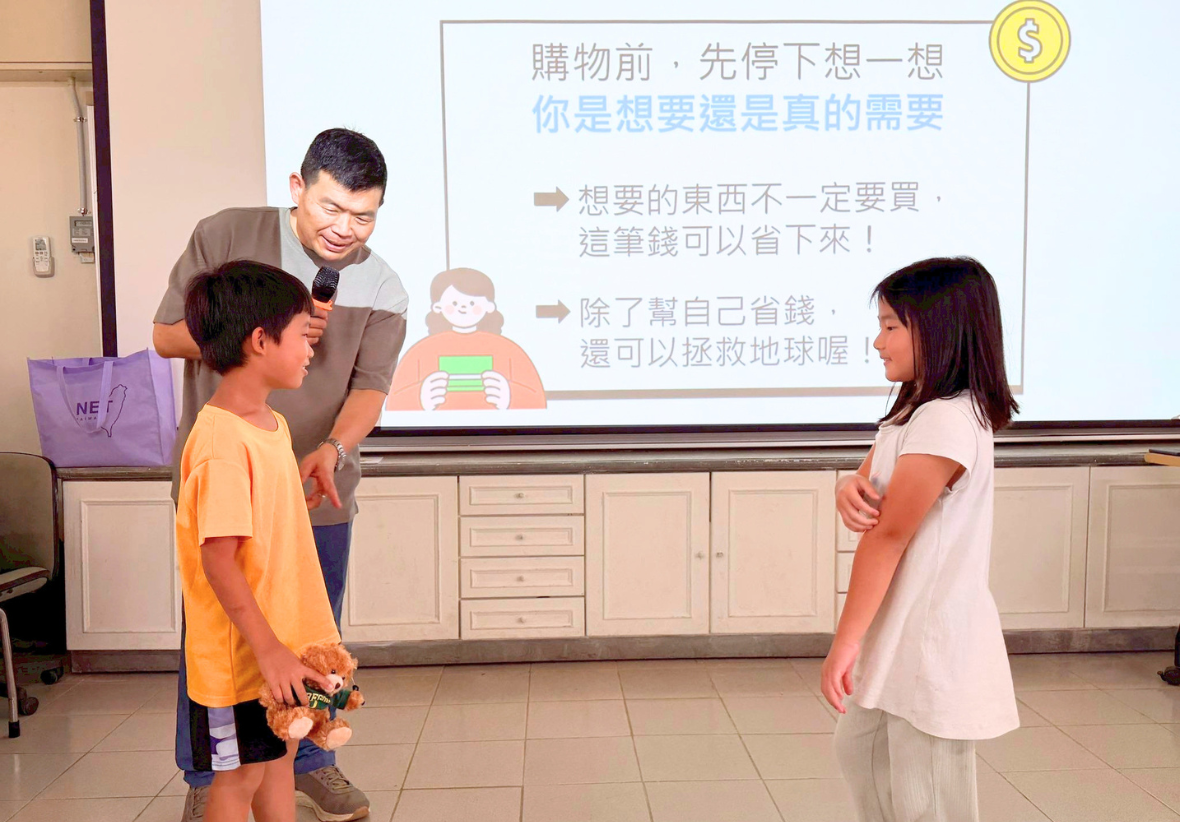 照片中，一位男講師手持麥克風，正在台上與一男一女兩名學童進行互動。男學童手裡拿著一隻泰迪熊玩偶。背景的投影螢幕上顯示著「購物前，先停下想一想：你是想要還是真的需要」的課程內容，以及兩點關於省錢和環保的說明