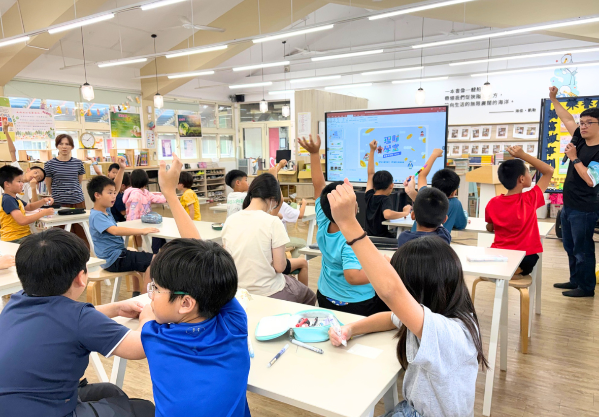 這是一張室內全景照片，拍攝於一個明亮的圖書館或多功能教室。許多國小學童坐在桌前，紛紛舉手，積極回答教室前方一名穿黑T恤、持麥克風的男講師的問題。投影螢幕上顯示著「互動學堂」的字樣。背景書架上擺滿了書籍，環境溫馨。