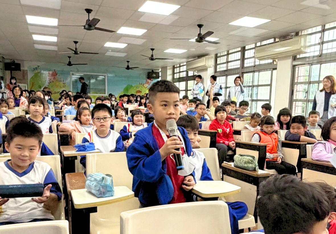 這是一張近攝照片，拍攝於階梯教室。一名穿藍色外套的男學童站在課桌椅間，雙手握著一支麥克風，神情認真地準備回答問題。他周圍坐滿了其他穿著制服的學童，許多人正看著他。背景可見其他工作人員和教室的窗戶，現場氣氛活潑。