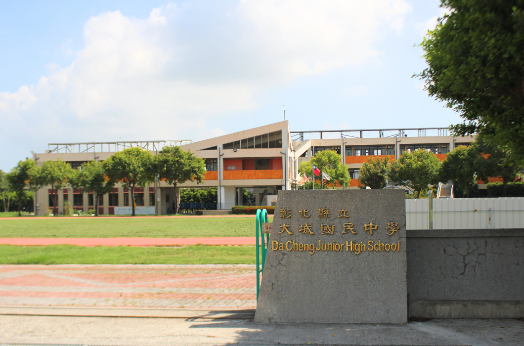 這張圖是彰化縣立大城國民中學的校門口外觀。前方立有一塊刻有金字校名的灰色石碑，後方可見紅磚色與米色相間的學校主建築物、紅色的操場跑道、草皮，以及一排綠意盎然的樹木。