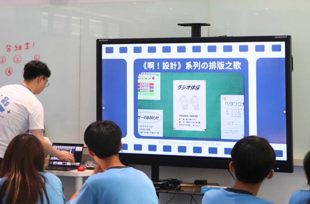 這是一張起駛簡報講師操作電腦的教學場景。大螢幕上顯示著日本《啊！設計》系列的排版之歌影片畫面。白衣男講師正彎腰操作桌上的筆記型電腦，台下幾位身穿藍色上衣的學生背對鏡頭觀看。