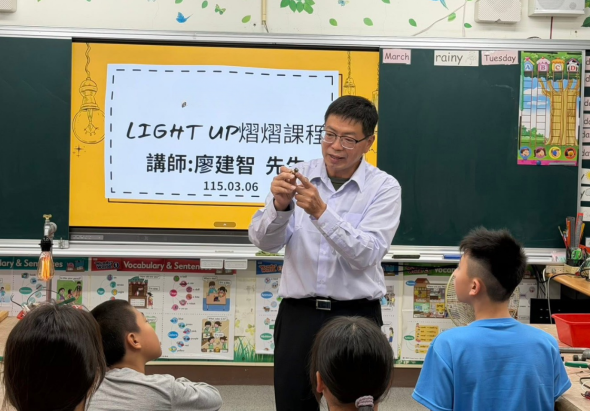 廖建智老師站在台前，手持小零件向台下的國小學生們講解。後方的黃色電子白板投影著「LIGHT UP 熠熠課程 講師：廖建智先生 115.03.06」字樣。