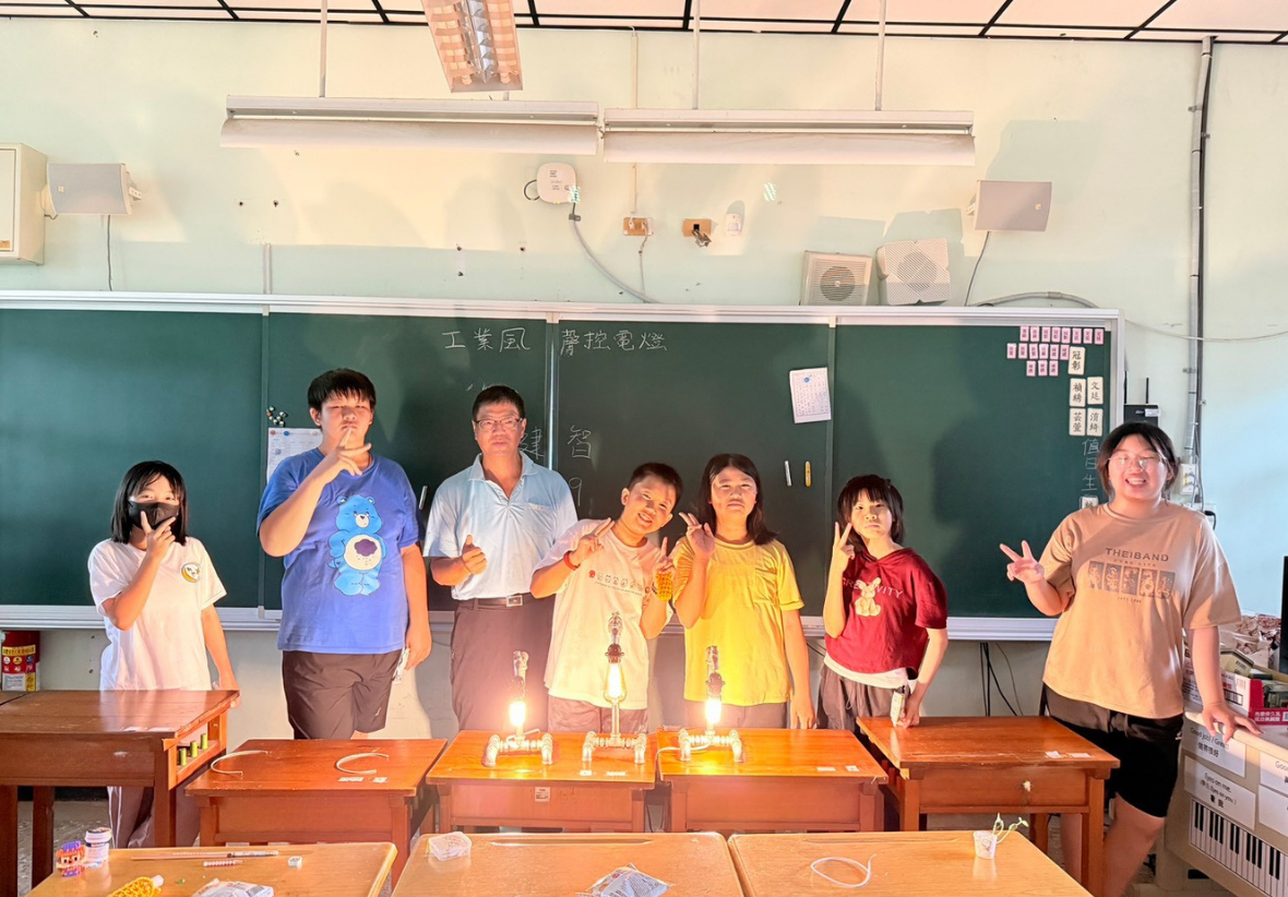 廖建智老師與六位國小學生在教室黑板前的合照。講台上放置著三盞發著暖黃光的水管造型工業風電燈，大家開心地面對鏡頭比出讚或剪刀手。