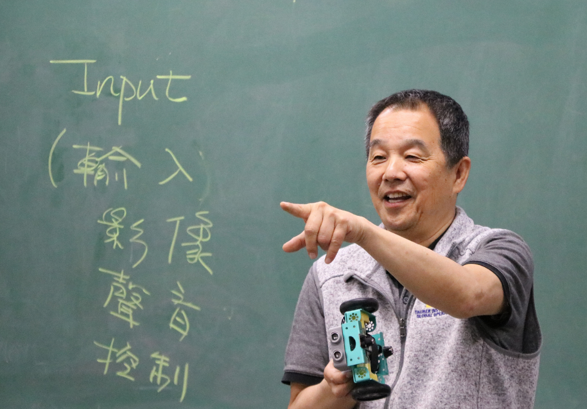 一位中年男性講師在綠色黑板前授課。黑板上寫著「Input (輸入)、影像、聲音、控制」，講師右手拿著一台教學用的小型輪式機器人模型，正微笑向前方指引。